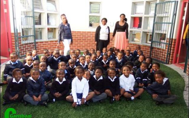 Die graad R-leerders sit hier op die sintetiese grasperk tussen die graad R-klaskamers met die argitektoniese voorkoms van luukse rondawels. Agter van links staan die opvoeders Xoliswa Hoza (Gr Ra), Ellen Pato (Gr Rb) en Monica Sam (Gr Rc).