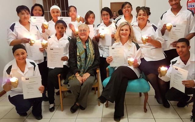 Die tuisversorgers met hul brandende lampies – voor van links: Celeste Dunsdon, Sanna Speelman (beste prestasie: strykwerk), Suster Irene de Waal, El-Marie Fourie (hoogste lof vir teorie en prakties) en Franklin Eksteen (eervolle vermelding).  Agter: Corné Dunsdon (beste blommerangskikking), Trudie Cupedo, Elna Kondokter (prakties en huishouding met lof), Rhowina Kammies, Magdalena Arends (naaldwerktoekenning), Nicole Adonis, Jocoline Louw (slaag prakties met lof), Marlene Gertse (klaskaptein – slaag prakties en tuisversorgingsteorie met lof) en Peter-Lee Karelse (slaag prakties met lof).